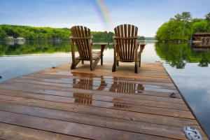 Two wooden chairs sit on a wet dock overlooking a calm lake, framed by lush green trees. The sky is clear with a faint rainbow reflected in the water, creating a serene, natural setting—a peaceful contrast to the fast-paced world of government just beyond this tranquil escape.