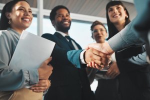 A group of diverse professionals in business attire stands proudly, exuding a sense of effective leadership. Two individuals in the foreground are shaking hands, leading the charge towards a successful agreement. The bright indoor setting with modern decor enhances their collaborative spirit.