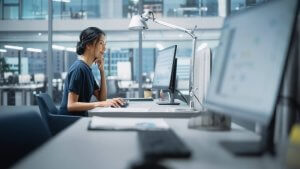 A woman sits at a desk in a modern office, looking at a computer monitor with graphs displayed. She appears focused, embodying the determination and insight often seen in leaders, with one hand on the mouse and the other gently touching her chin.