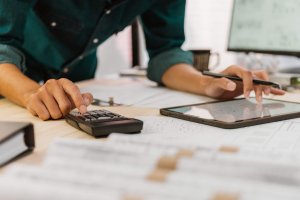 A person at a busy workplace uses a calculator and digital tablet at a desk cluttered with documents, analyzing financial data or handling accounting tasks.