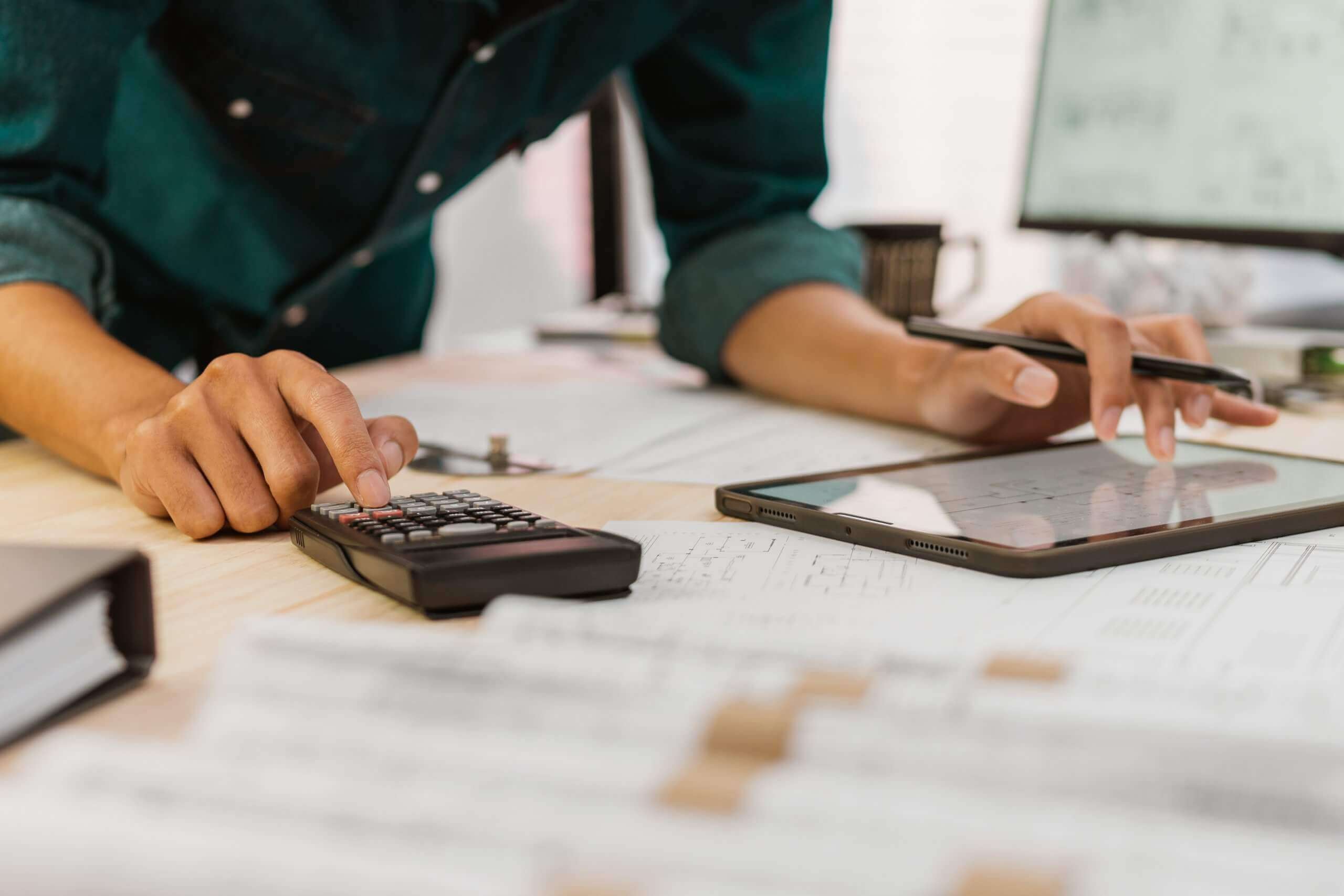 An employee using a calculator and holding a pen, working at a desk covered with documents, a binder, and a tablet, with a computer monitor in the background. An employee using a calculator and holding a pen, working at a desk covered with documents, a binder, and a tablet, with a computer monitor in the background.