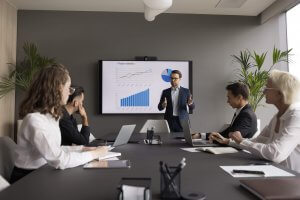 A man in a suit demonstrates leadership as he presents charts and graphs to four colleagues seated around a conference table in a modern office filled with laptops and notebooks.