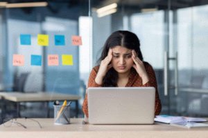 A woman sits at a desk with her laptop, looking stressed and disengaged as she holds her head in her hands. Sticky notes cover the glass wall behind her, while papers and a pencil holder clutter the desk.