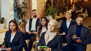 A diverse group of young adults in business attire sit indoors, attentively taking notes during a DEI-focused seminar or workshop, with notebooks and pens in hand.