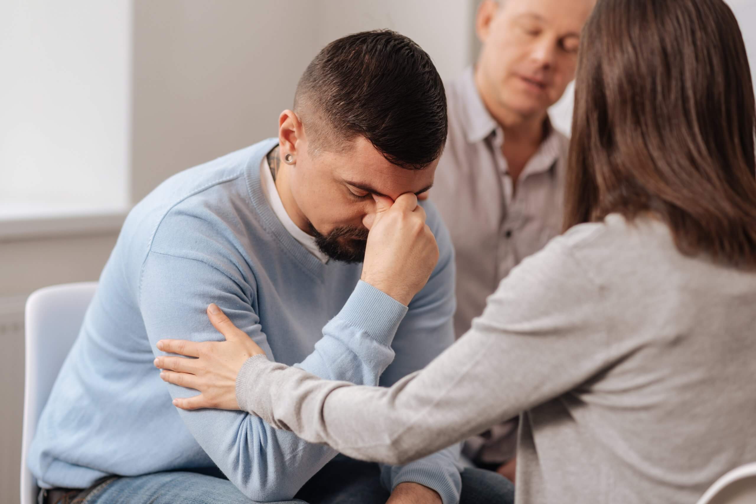A man sits with his head bowed and hand covering his face, looking distressed. A woman in the foreground gently touches his arm for support, while another man sits nearby, watching with concern in this DEI-focused moment of empathy.