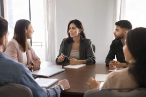 Five leaders sit around a conference table, engaged in discussion. A woman in a blazer speaks while others listen attentively, with notebooks and laptops in front of them. The room is bright with natural light from windows.
