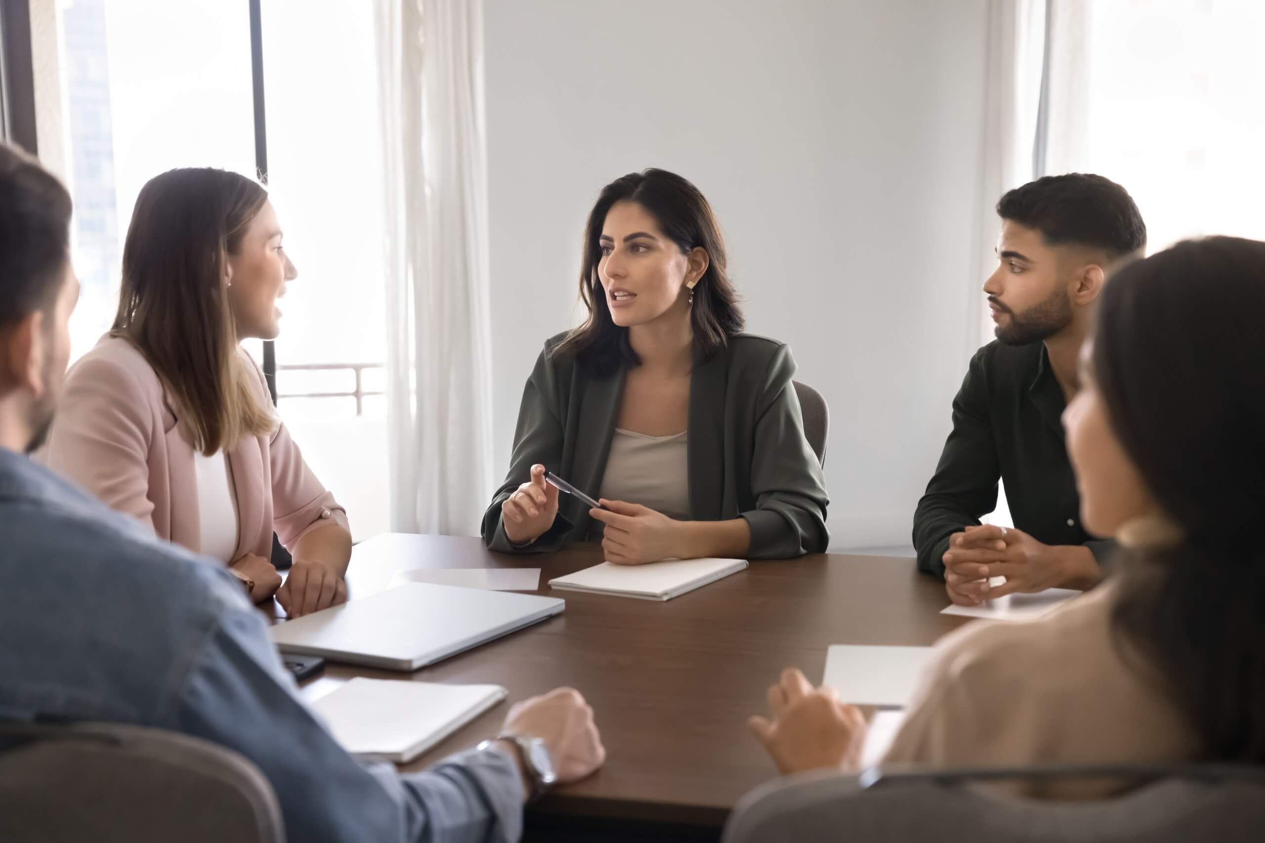 Five team members sit around a conference table in discussion. A woman in the center speaks while others listen attentively, with notebooks and laptops in front of them. Natural light comes through windows in the background.