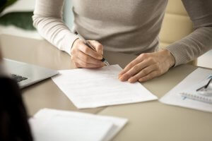 A person wearing a light sweater is sitting at a workplace desk, holding a pen and filling out or signing a document. Papers and a laptop are visible on the desk.