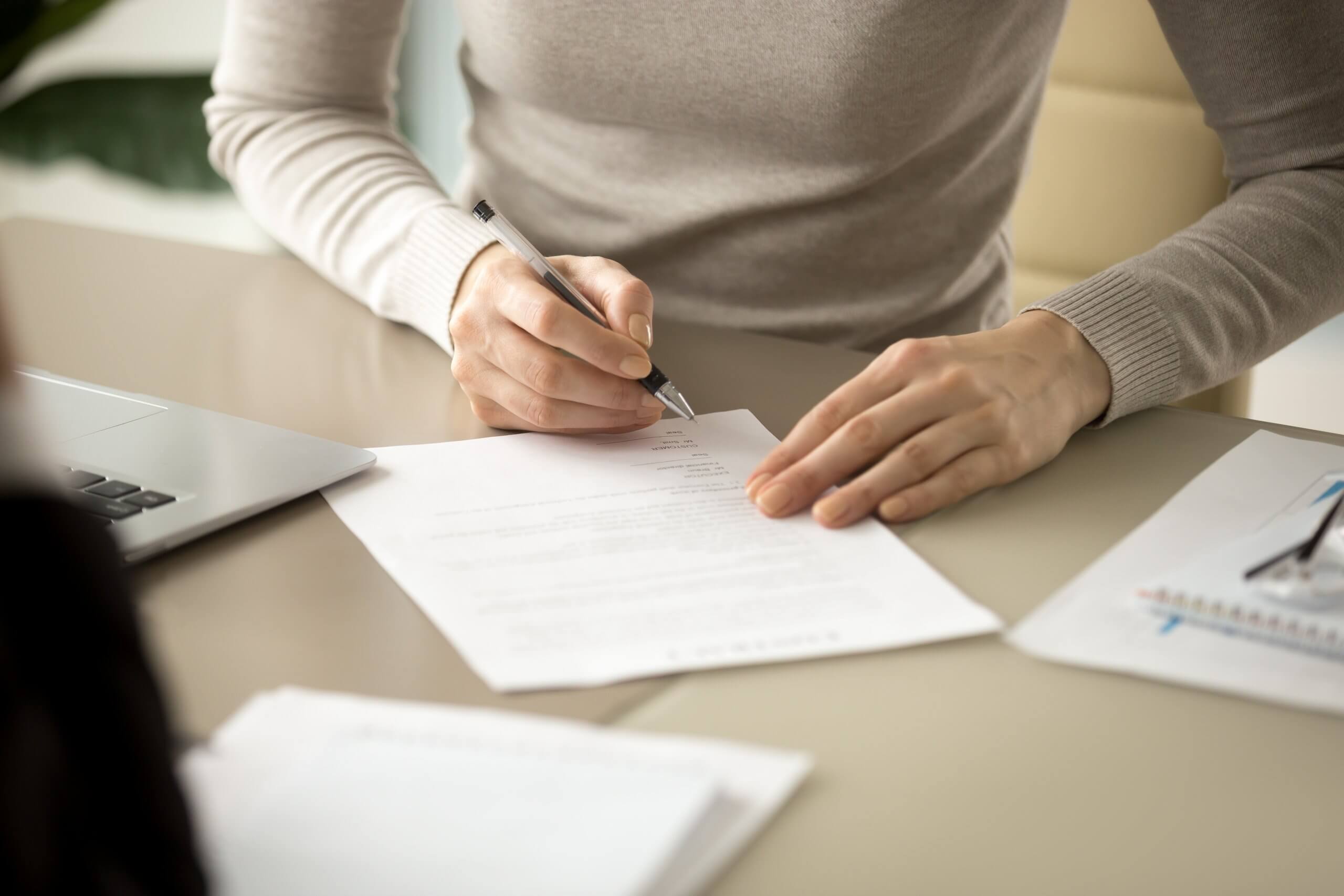 Close-up of a person in a light sweater sitting at a workplace desk, holding a pen and filling out or signing a document, with a laptop and notepad nearby. Close-up of a person in a light sweater sitting at a workplace desk, holding a pen and filling out or signing a document, with a laptop and notepad nearby.