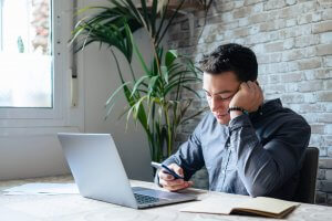A man sits at a desk with his laptop, looking at his phone and smiling. An open notebook is in front of him, yet signs of disengagement are clear as he’s focused on his phone instead of work. A plant and a brick wall are in the background.