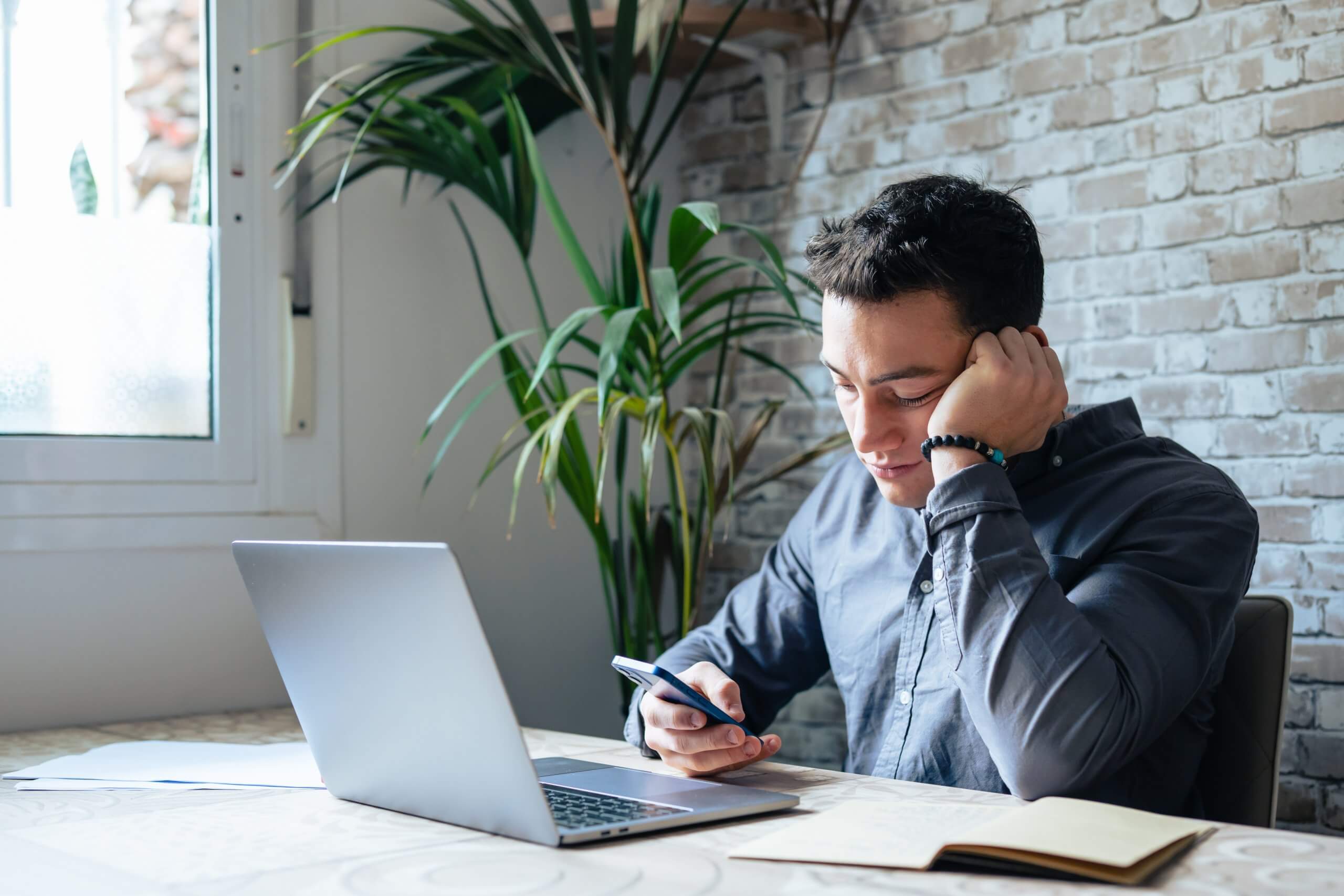 A man sits at a table with a laptop, smiling at his smartphone as he enjoys conversations. Indoors by a window and a large plant, he’s surrounded by notebooks and papers on the table.