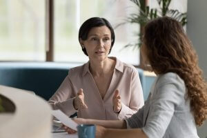 Two women sit at a table in an office, having a serious conversation. One woman gestures with her hands while speaking; the other listens, holding a document and a blue mug. Employers' discussions like these can shape decisions. Green plants are visible in the background.