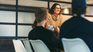 A woman wearing glasses and a sweater smiles while leading a casual, friendly conversation with two people seated at a table in a modern office setting. One person holds a cup, adding to the warm atmosphere.