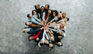 A diverse group of people standing in a circle, symbolizing leadership as they reach into the center and place their hands together, photographed from above on a gray background.