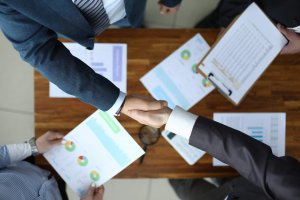 Two people in business attire shake hands over a wooden table with documents and charts, while two others hold papers and a clipboard, illustrating a professional workplace meeting or agreement.