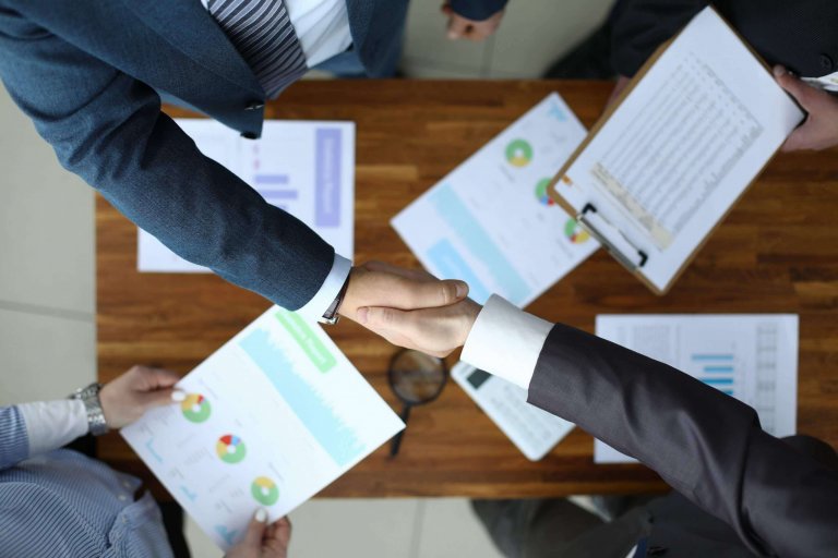 Two people in business attire shake hands over a wooden table with documents and charts, while two others hold papers and a clipboard, illustrating a professional workplace meeting or agreement.