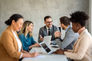 Five people sit around a conference table, engaged in a lively discussion. Two leaders are focused on laptops, while others are talking and taking notes. Everyone appears to be smiling and collaborating in an office setting.