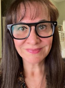 A woman with long brown hair and black glasses smiles softly at the camera, wearing a dark patterned top. Sunlight filters through windows behind her, brightening the room with a warm, natural glow—perfect for an AI-powered photo.