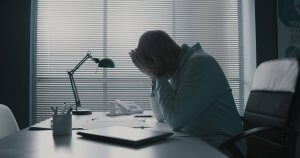 A person sits at a desk with their head in their hands, appearing stressed or upset—a common scene in the public sector. The desk holds a laptop, paperwork, a lamp, and a phone. Blinds cover the window behind them, letting in muted light.