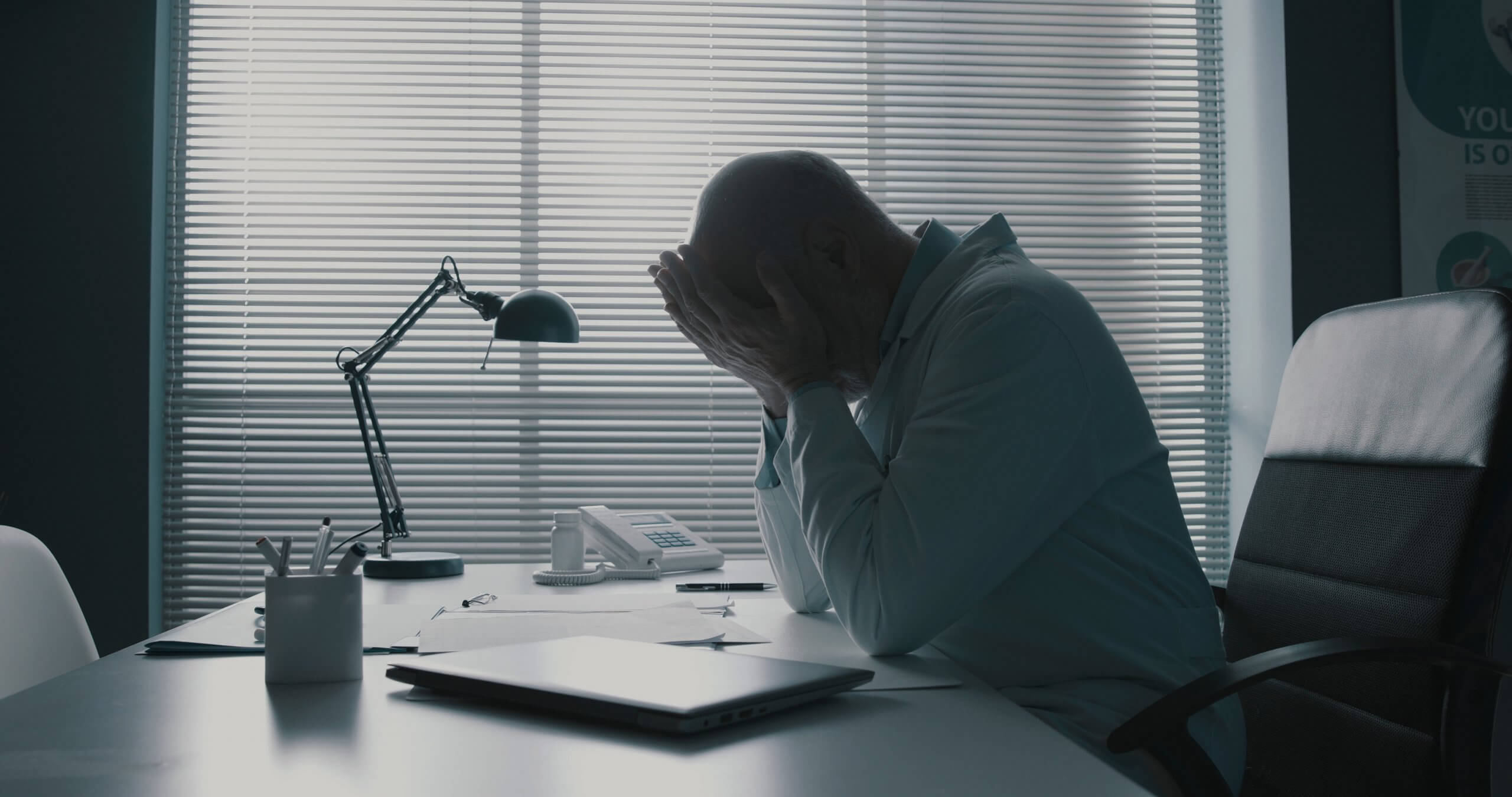 A person in a lab coat sits at a desk with their head in their hands, appearing stressed, possibly over government regulations. The room is dimly lit, with closed blinds letting in some light behind them. A person in a lab coat sits at a desk with their head in their hands, appearing stressed, possibly over government regulations. The room is dimly lit, with closed blinds letting in some light behind them.