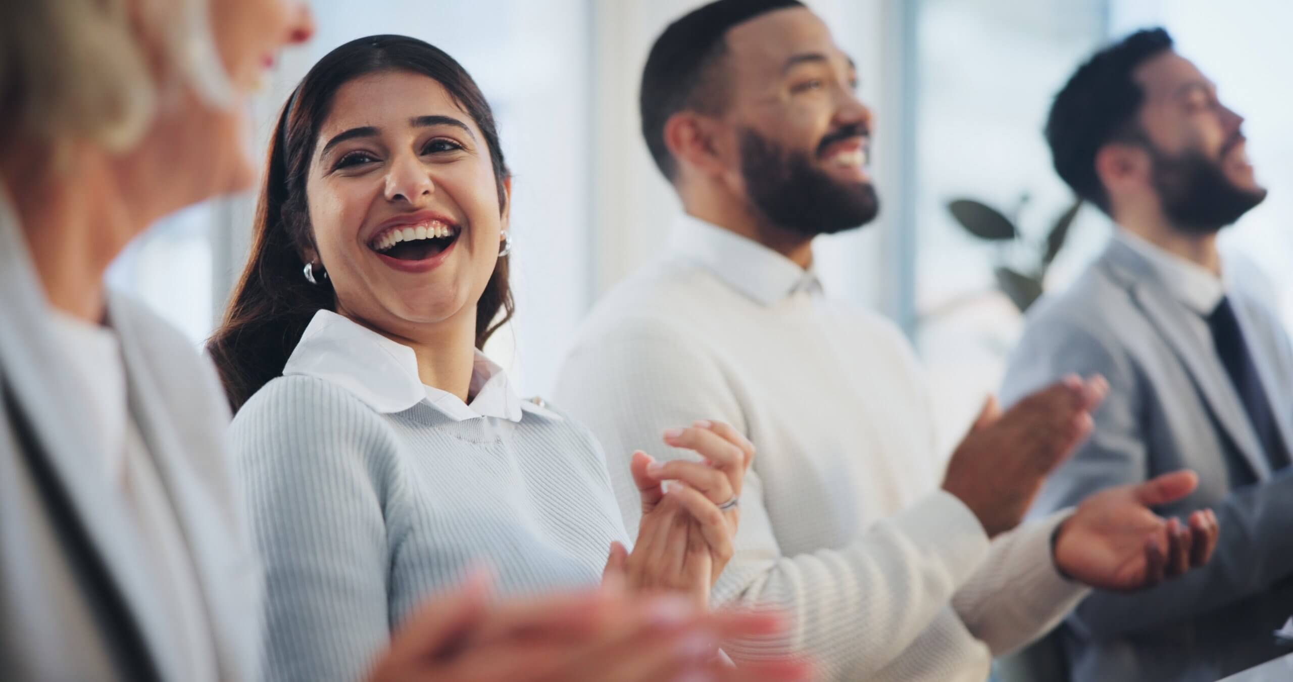 A group of people in business attire smiling and clapping, exuding a sense of engagement and happiness in a bright indoor setting. A group of people in business attire smiling and clapping, exuding a sense of engagement and happiness in a bright indoor setting.