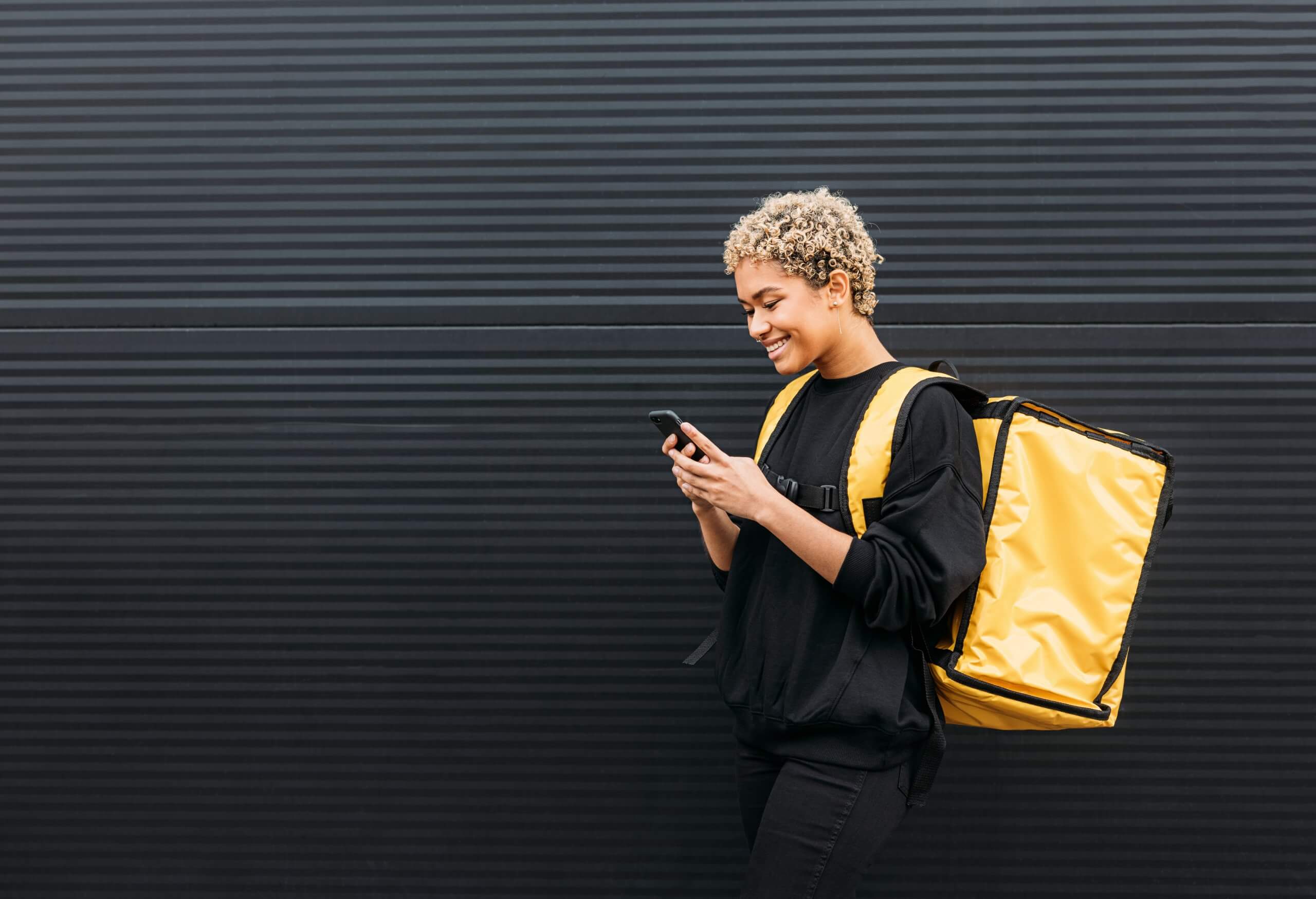 A person wearing a black outfit and large yellow insulated backpack smiles while using their phone, perhaps texting employers, as they stand in front of a dark, textured wall. A person wearing a black outfit and large yellow insulated backpack smiles while using their phone, perhaps texting employers, as they stand in front of a dark, textured wall.