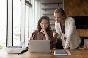 Two women in an office sit at a wooden table looking at a laptop. One woman sits thoughtfully with her hand on her chin, while the other leans over, pointing at the screen and discussing career opportunities.