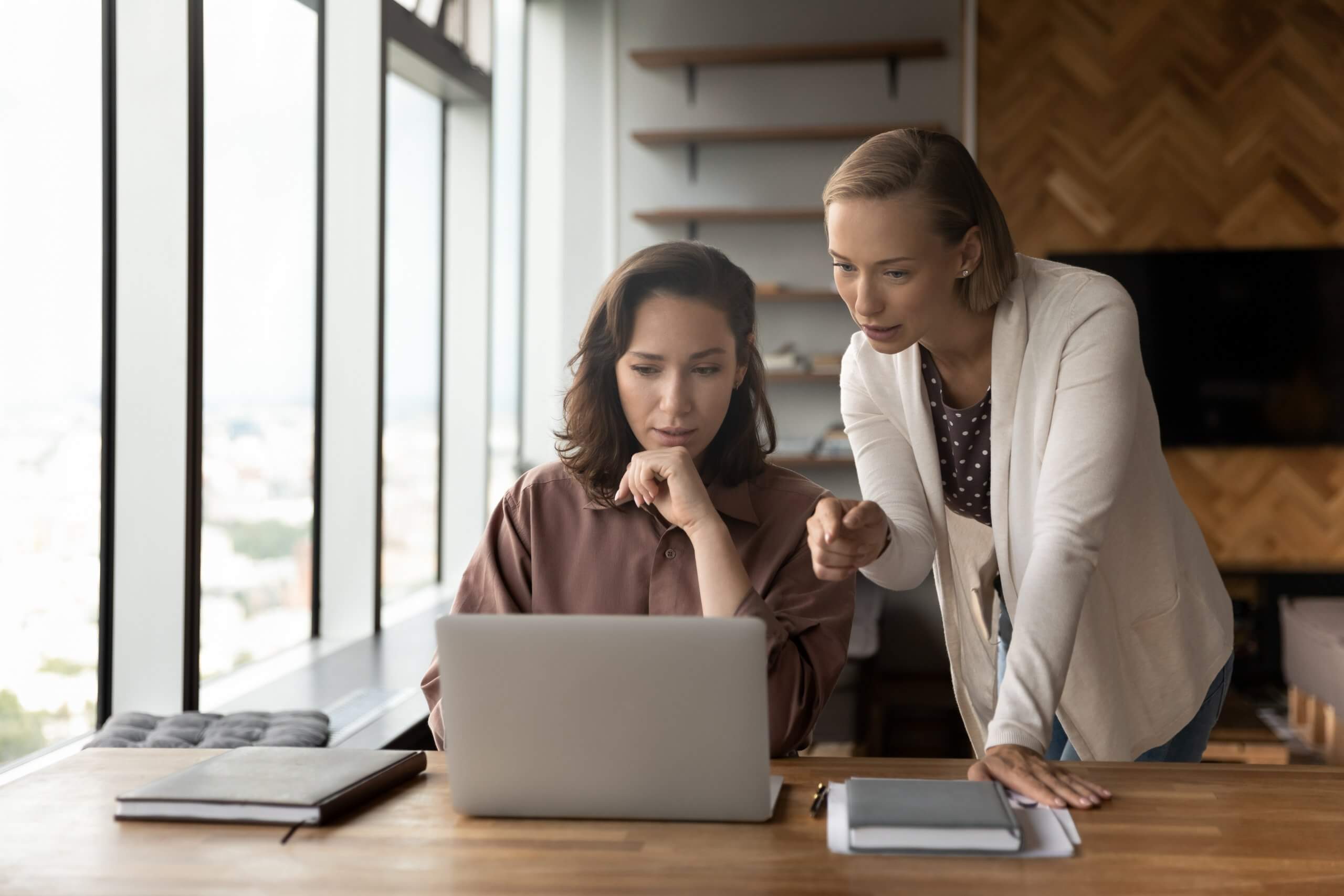 Two women in a government office look at a laptop together; one is seated and focused on the screen, while the other stands beside her, pointing at the laptop and explaining something. Large windows let in natural light. Two women in a government office look at a laptop together; one is seated and focused on the screen, while the other stands beside her, pointing at the laptop and explaining something. Large windows let in natural light.