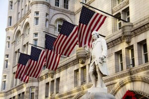 A white marble statue stands in front of a historic stone building, symbolizing civil service, with four large American flags displayed in a row along the building’s facade.