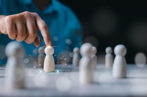 A hand points to a glowing wooden figure among several plain figures on a table, symbolizing leadership, selection, or individuality with a blue background.