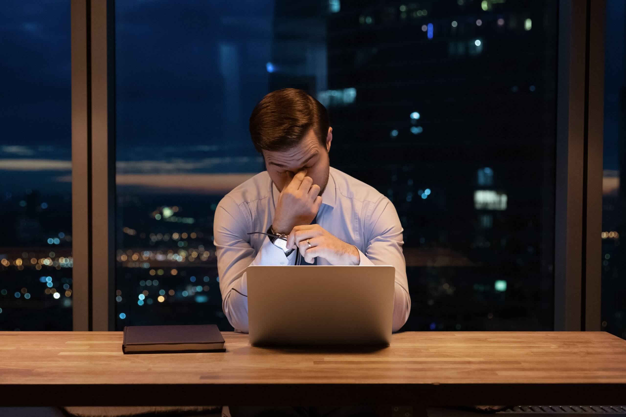 A man in a white shirt sits at a desk with a laptop, pinching the bridge of his nose in frustration. A closed book lies nearby, while the cityscape glows at night—perhaps the stress of government work weighing on his mind. A man in a white shirt sits at a desk with a laptop, pinching the bridge of his nose in frustration. A closed book lies nearby, while the cityscape glows at night—perhaps the stress of government work weighing on his mind.