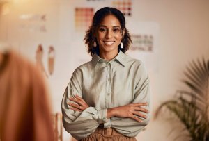 A woman with curly hair smiles confidently with her arms crossed, embodying success in a light-colored blouse and earrings, standing indoors in a warmly lit room with blurred charts and plants in the background.