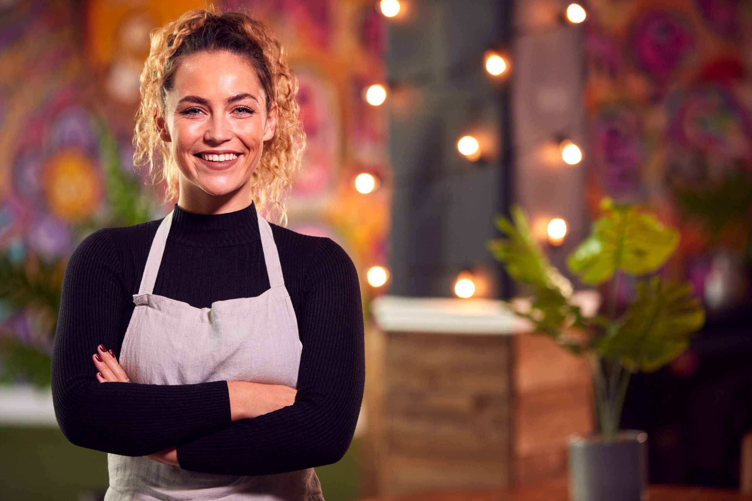 A smiling woman wearing a light-colored apron stands with arms crossed in a colorful café or restaurant, where creative employers have adorned the space with string lights and vibrant wall art in the background. A smiling woman wearing a light-colored apron stands with arms crossed in a colorful café or restaurant, where creative employers have adorned the space with string lights and vibrant wall art in the background.