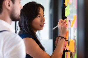 A woman demonstrates leadership as she writes on a sticky note attached to a glass wall filled with colorful notes, while a man stands nearby, observing her work.