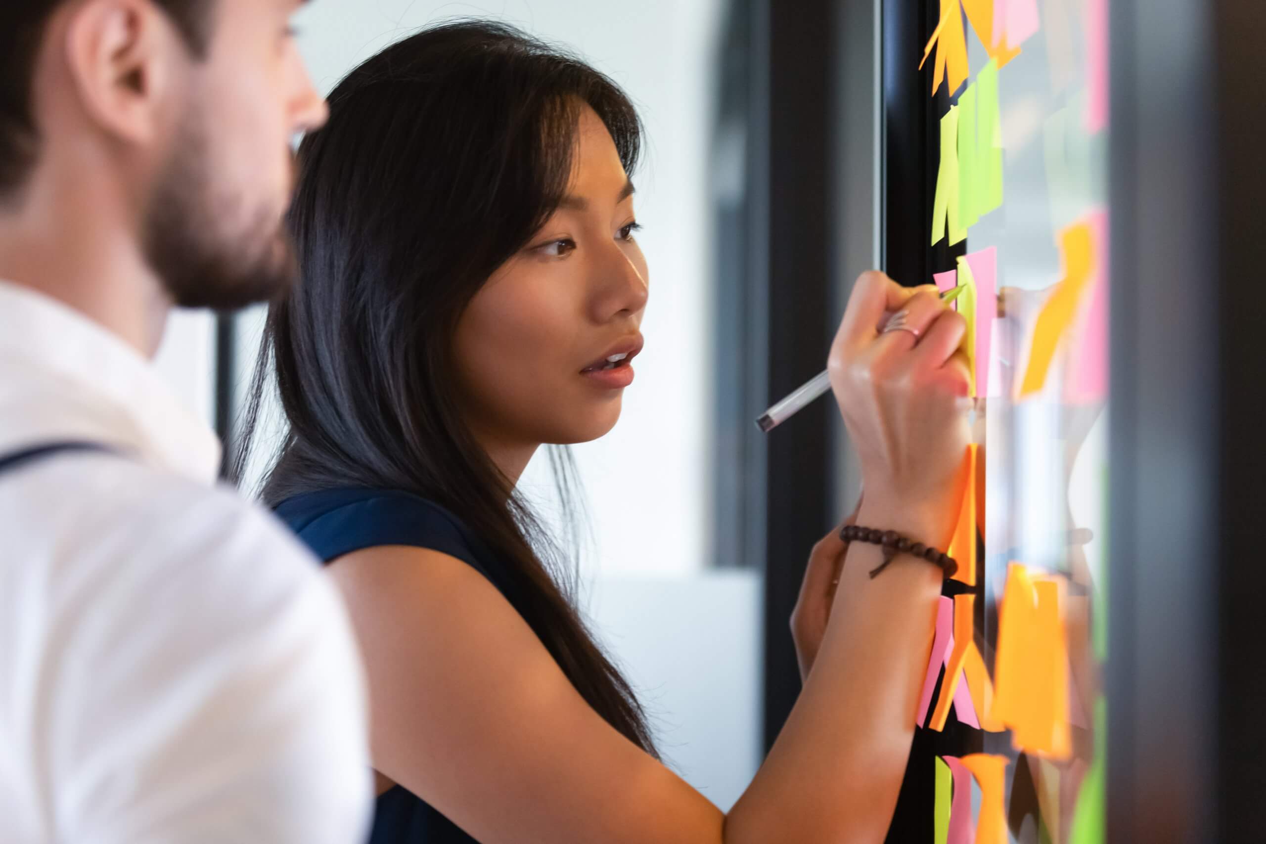A woman writes on a transparent board covered with colorful sticky notes, while a man stands nearby watching. Their active collaboration in the modern office contrasts sharply with workplace disengagement. A woman writes on a transparent board covered with colorful sticky notes, while a man stands nearby watching. Their active collaboration in the modern office contrasts sharply with workplace disengagement.