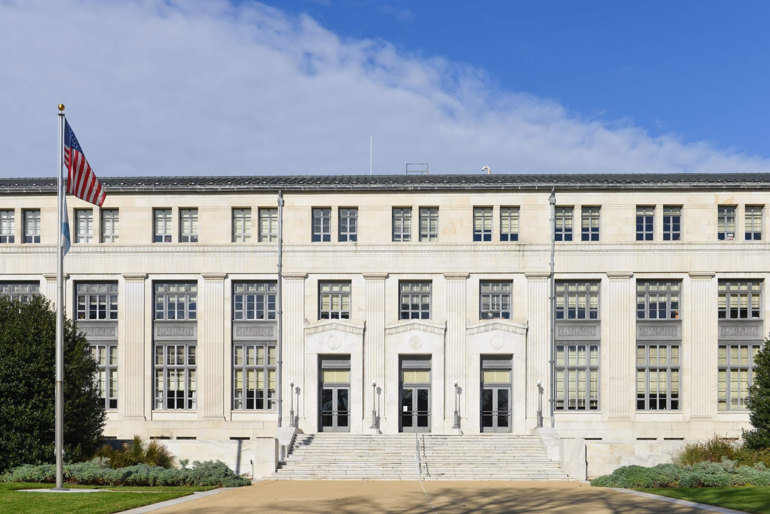 A large, three-story government building with a symmetrical facade, tall windows, and double entrance doors. Stairs lead up to the entrance. An American flag is on a pole in front, under a partly cloudy sky. A large, three-story government building with a symmetrical facade, tall windows, and double entrance doors. Stairs lead up to the entrance. An American flag is on a pole in front, under a partly cloudy sky.