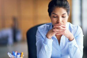 A woman in a light blue shirt sits at a desk, hands clasped in front of her mouth, looking down thoughtfully—perhaps reflecting on the challenges faced in the public sector.