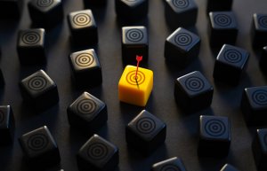 A yellow cube with a target symbol and a red arrow in its center stands out among many black cubes with target symbols on a dark surface, creating a sense of focus and direction.
