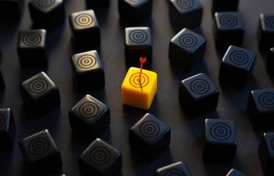 A group of black cubes with target symbols surround a single yellow cube, marked for success, which has a small red arrow stuck in its center. The yellow cube stands out among the black ones.