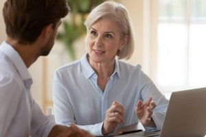 An older woman with short gray hair is talking to a younger man about his career while sitting at a table with a laptop. She appears engaged and expressive, using her hands while speaking.