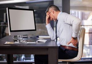 A man in business attire sits at a desk in a government office, holding his lower back with one hand and his forehead with the other, appearing to be in pain or discomfort while working at a computer.