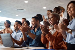 A diverse group of people sitting together indoors, smiling and applauding enthusiastically, suggesting they are celebrating or acknowledging a collaborative achievement. A laptop and documents are visible in the foreground.
