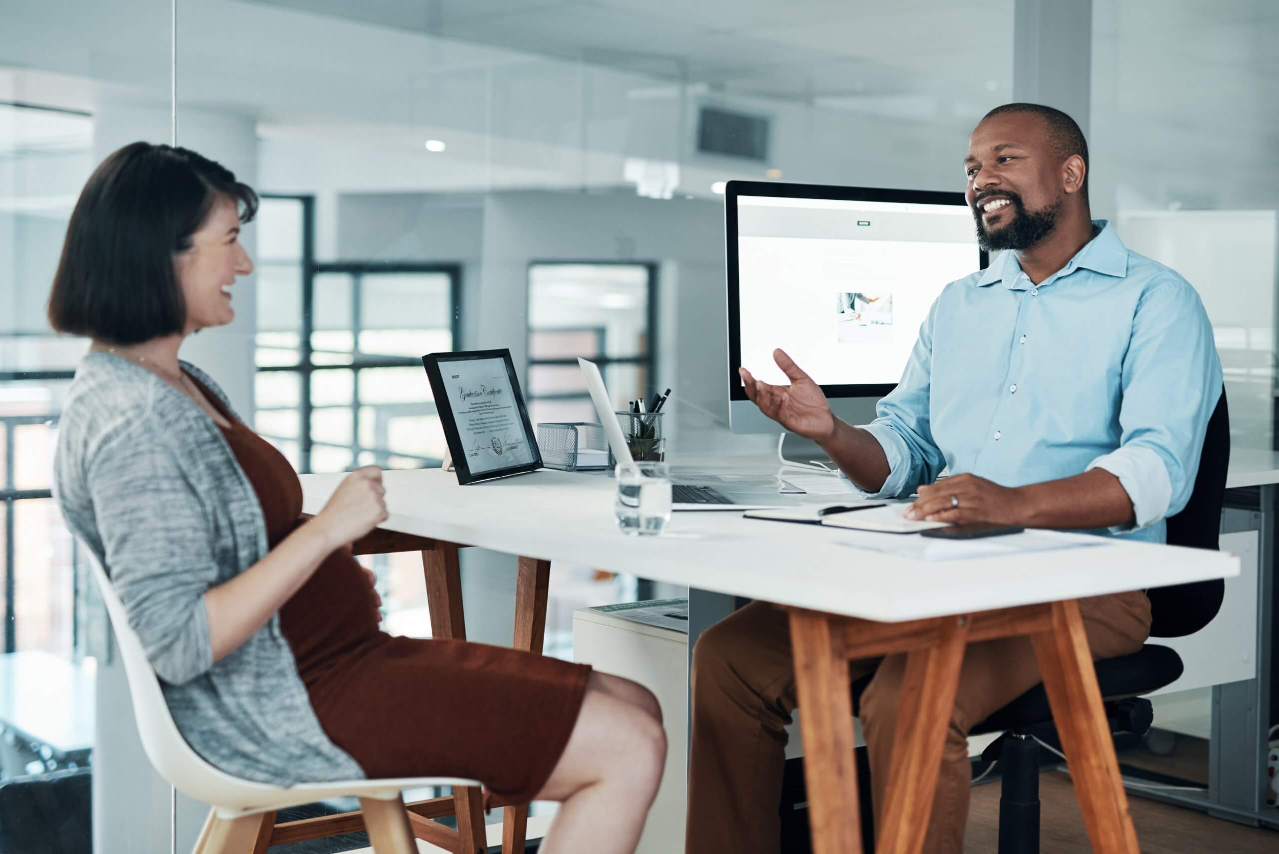A man and a woman sit across from each other at an office desk, smiling and engaged in conversation as they learn from one another. The desk holds a tablet, papers, and a glass of water, set against a bright, modern office space. A man and a woman sit across from each other at an office desk, smiling and engaged in conversation as they learn from one another. The desk holds a tablet, papers, and a glass of water, set against a bright, modern office space.