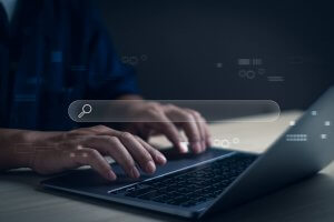 Person typing on a laptop with a transparent search bar and search icon superimposed, suggesting an online job search or internet browsing concept. The background is dark and the focus is on the hands and keyboard.