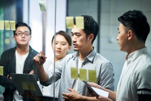 Four employees work together in an office, standing around a glass board with sticky notes. One man writes or draws on the board as the others look on, holding a laptop and papers, appearing engaged and focused.
