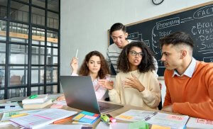 Four young adults sit and stand around a laptop at a table covered with papers, notebooks, and stationery. One gestures while discussing governments, as a chalkboard with diagrams is visible in the background.