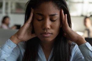 A young woman with closed eyes holds her hands to her temples, appearing stressed or overwhelmed by government concerns, while sitting indoors. Other people are blurred in the background.