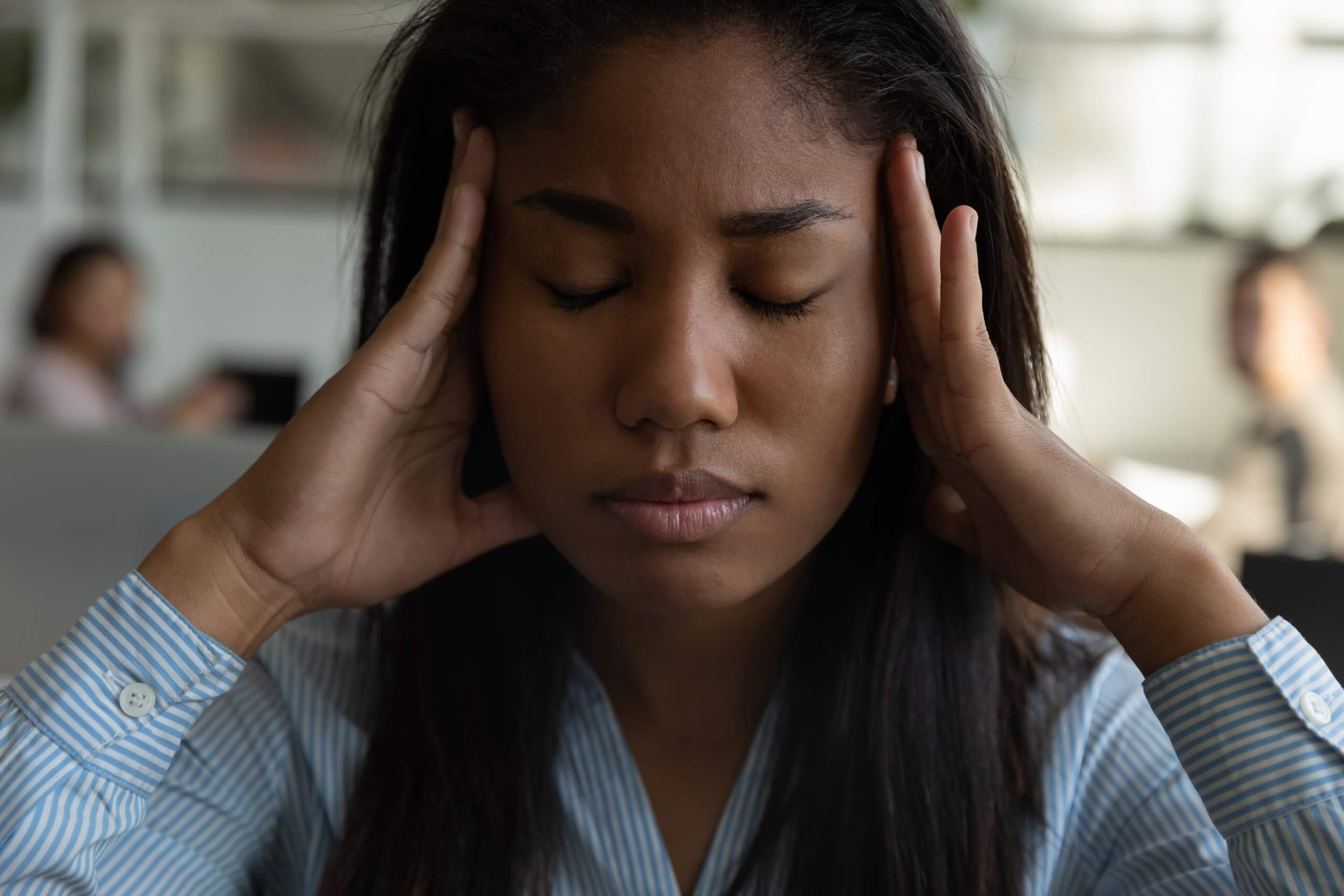 A woman with closed eyes presses her hands to her temples, appearing stressed or in pain, possibly due to performance management pressures, while sitting indoors. The blurred background reveals other people in an office setting. A woman with closed eyes presses her hands to her temples, appearing stressed or in pain, possibly due to performance management pressures, while sitting indoors. The blurred background reveals other people in an office setting.
