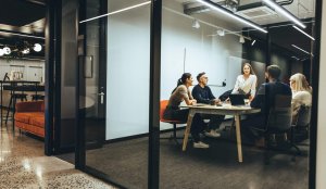 A group of six people sit around a table in a modern glass-walled office, engaged in a collaboration-focused meeting. One woman stands and speaks while others listen, with laptops and papers spread across the table.
