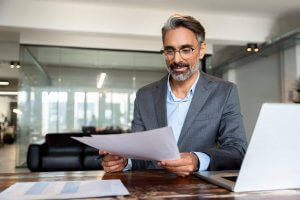 A man in a gray suit with glasses and gray hair sits at a desk, reviewing performance papers. A laptop and additional documents rest on the wooden table in a modern office setting with glass walls in the background.
